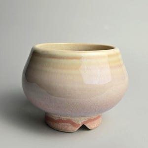 Peut inclure: Un bol en céramique avec un corps arrondi et une base sur pied. L'émail présente une transition en dégradé allant d'un blanc cassé crémeux sur le bord à une teinte rose et lavande douce vers le bas. La base montre une bande horizontale rouge-brun.