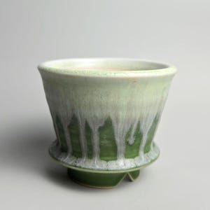 May include: A ceramic cup featuring a flared rim and a footed base. The exterior displays a two-tone glaze with a deep forest green bottom transitioning into a lighter mint green top, characterised by white glaze drips running down the sides.