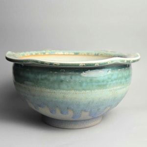 May include: A ceramic bowl with a scalloped rim, featuring a gradient glaze that transitions from a deep teal at the top to a lighter blue and off-white base. The exterior surface displays a mottled texture with visible drip patterns.