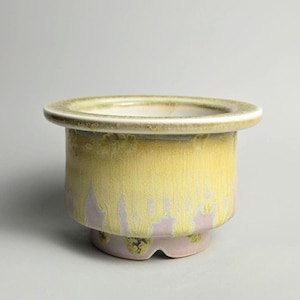 May include: A ceramic planter with a wide, circular rim and a cylindrical body. The planter features a yellow glaze that drips down over a lavender base. The rim is a lighter shade of yellow. The planter is designed to hold plants.