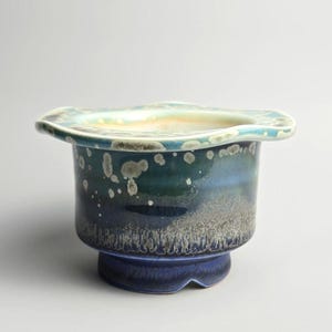 Puede incluir: Una maceta de cerámica hecha a mano con una forma orgánica única. La maceta presenta una base azul oscuro, que transiciona a tonos de verde y turquesa, con acentos moteados. El borde tiene un borde ondulado y un color azul claro y crema.