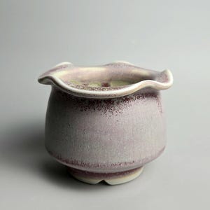 May include: A small ceramic planter with a rounded body and a ruffled, wavy rim. The exterior features a mottled glaze with shades of dusty lavender, soft pink, and deep burgundy speckles, transitioning to a cream-coloured base and interior.