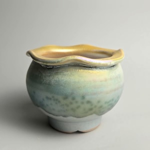 May include: A small ceramic bowl with a rounded body and a wavy, scalloped rim. The exterior features a gradient glaze transitioning from a pale, light blue at the base to a soft, muted green in the centre, topped with a warm, golden yellow edge.