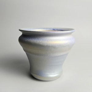 May include: A ceramic planter with a flared rim and a rounded body. The surface features a pearlescent, iridescent glaze that shifts between soft shades of light blue, lavender, and subtle gold, creating a shimmering, reflective finish.
