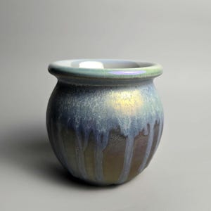 May include: A small ceramic pot with a rounded body and a wide, flared rim. The exterior features a reactive glaze with a colour gradient transitioning from a pale, iridescent mint green at the top to a deep, mottled blue and earthy brown base.