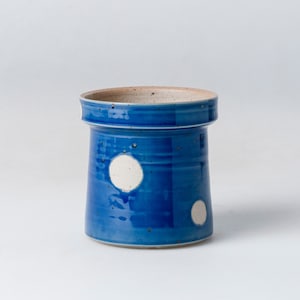 Peut inclure: Un pot à ustensiles en céramique bleu avec des pois blancs. Le récipient cylindrique a un large bord et un intérieur moucheté non émaillé. L'extérieur est bleu vif avec une finition brillante, mettant en valeur la qualité artisanale de la poterie.