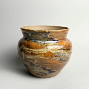 May include: A ceramic pot with a wide, flared rim and a rounded body. The exterior features a variegated glaze in shades of amber, brown, and cream, with streaks of metallic blue and grey accents layered across the textured, glossy surface.