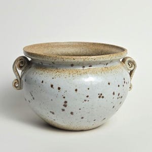 May include: A white ceramic planter with brown speckles and two curled handles. The planter has a rounded shape and a slightly flared rim.