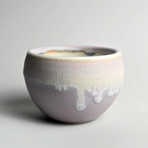Puede incluir: Cuenco de cerámica hecho a mano con forma redondeada. El cuenco presenta un degradado de color, pasando de un crema claro en el borde a un lavanda suave en la base. El esmalte tiene un efecto goteado, añadiendo textura.