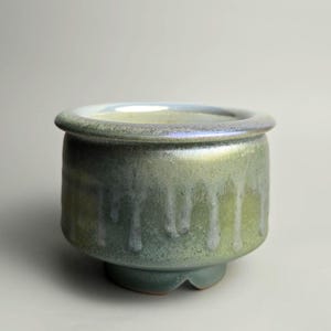 May include: A small ceramic bowl with a rounded body and a wide, flat rim. The exterior features a variegated glaze in shades of sage green, olive, and soft blue, with a distinct drip pattern running vertically down the sides toward the base.