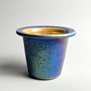 May include: A small, handcrafted ceramic pot with a flared rim. The exterior has a textured, iridescent finish in shades of blue and green, while the interior and rim are gold. The pot is approximately 7.6 cm tall.