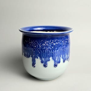 May include: A ceramic plant pot with a rounded form. The top rim is a deep blue, fading to a white base. The blue section has a drip design with white speckles, giving a textured look. Suitable for indoor or outdoor use. The pot is approximately 15 cm in height.