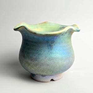 May include: A ceramic vase with a ruffled rim, displaying a mix of blue, green, and iridescent colours. The vase has a rounded body and a flared, wavy top, creating a unique and decorative piece.