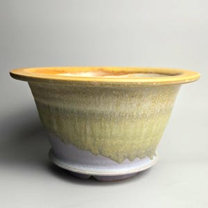 May include: A ceramic bowl with a wide, flat rim and a tapered body. The exterior features a textured, mottled glaze transitioning from a pale, dusty lavender at the base to a muted, earthy olive green and a warm, golden yellow along the top edge.