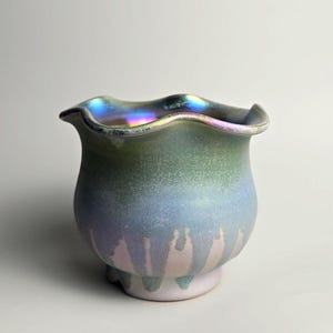 May include: A small, rounded glass bowl featuring an iridescent finish that transitions from a deep green at the ruffled rim to a soft blue and pale pink at the base. The surface has a shimmering, pearlescent quality with a wavy, scalloped edge.
