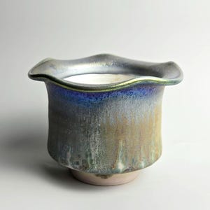 May include: A ceramic vase with a ruffled rim and a flared base. The vase has a metallic finish with shades of blue, green, and gold. The interior is white, and the base is a pale pink.