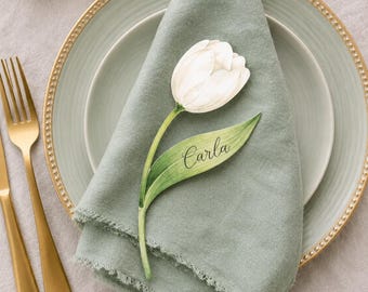 May include: A place setting with a gold-rimmed plate, a sage green napkin, and a white tulip-shaped place card that reads "Carla." Gold-colored fork and knife are on the left. The setting is on a light gray tablecloth.