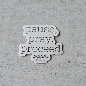 May include: White sticker with the words "pause, pray, proceed" in a black, handwritten-style font. The sticker also includes the text "teetotaler trinkets" in a smaller, cursive font. The sticker is on a light gray, textured surface.