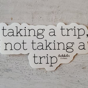 Può includere: Adesivo bianco con testo nero che recita "taking a trip, not taking a trip" con il testo "teetotaler trinkets" in un carattere più piccolo sotto il testo principale.