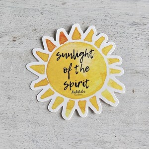 Può includere: Un adesivo fustellato a forma di sole con effetto acquerello. L'adesivo presenta le parole "sunlight of the spirit" in scrittura nera, con il nome dell'artista in basso. Il sole è giallo e arancione.