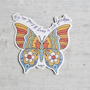 May include: A colorful butterfly sticker with a retro design. The sticker features a butterfly with orange, yellow, blue, and green wings. The text "We are going to know a new freedom" is written across the top. The sticker also includes the text "teetotaler trinkets".