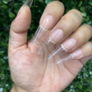 May include: Clear stiletto shaped false nails with a white tip. The nails are on a hand.