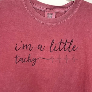 Pode incluir: Uma camiseta de cor vinho com o texto "i'm a little tachy" e um gráfico de batimento cardíaco estilizado.