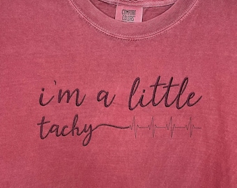 Può includere: Una maglietta color granata con il testo "i'm a little tachy" e una grafica di battito cardiaco stilizzata.
