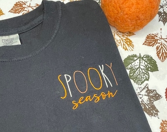 May include: Black t-shirt with the text "SPOOKY season" in orange embroidery on a white background. The shirt is lying on a white fabric with a brown and green leaf pattern. A small orange pumpkin is in the background.