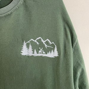 Pu&ograve; includere: Una maglietta verde con un disegno ricamato bianco di una catena montuosa con alberi e un cervo.