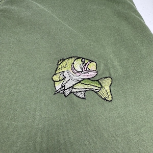 Può includere: Maglietta verde con un'immagine ricamata di un pesce persico.
