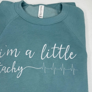 Può includere: Una felpa blu turchese con il testo "i'm a little tachy" e una linea ECG ricamata in bianco.