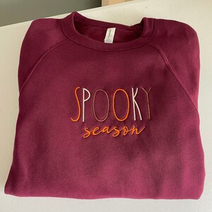 May include: Burgundy sweatshirt with the text "SPOOKY season" embroidered in white, orange, and brown thread.