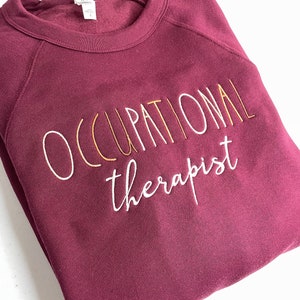 Może przedstawiać: Bordowa bluza z haftem "OCCUPATIONAL therapist" w kolorze białym i złotym.