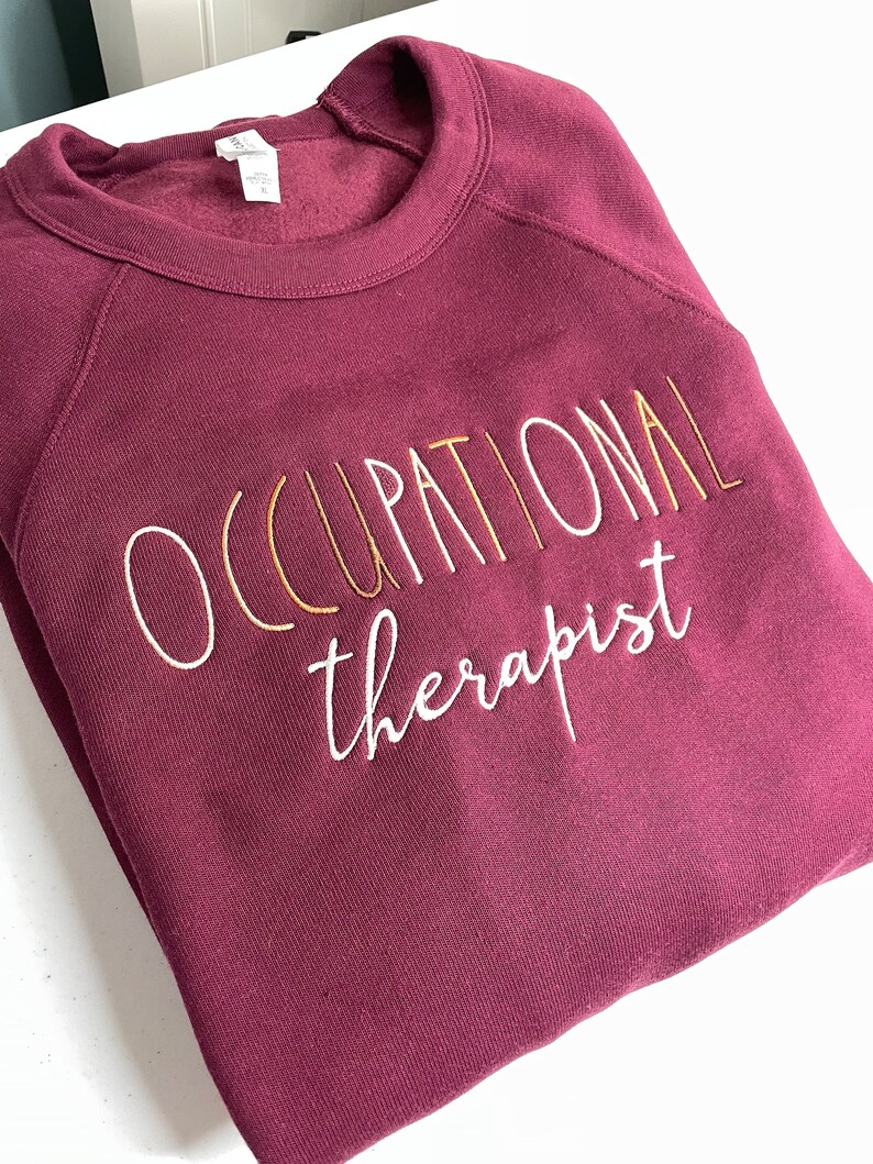 Może przedstawiać: Bordowa bluza z haftem "OCCUPATIONAL therapist" w kolorze białym i złotym.