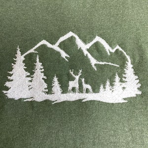 Pu&ograve; includere: Disegno ricamato bianco di una catena montuosa con due cervi davanti a una foresta su uno sfondo verde.