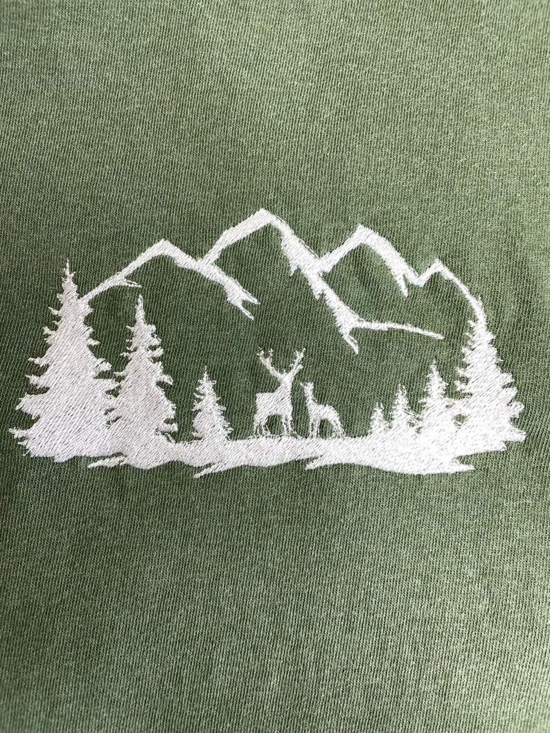 Pu&ograve; includere: Disegno ricamato bianco di una catena montuosa con due cervi davanti a una foresta su uno sfondo verde.
