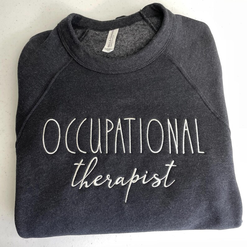 Może przedstawiać: Szara bluza z haftem "OCCUPATIONAL therapist" w kolorze białym.