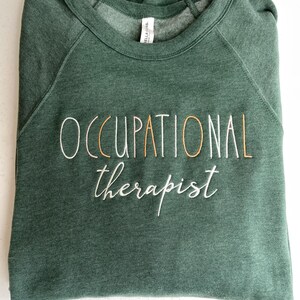 Może przedstawiać: Zielony sweter z haftem "OCCUPATIONAL therapist" w kolorze białym i złotym.
