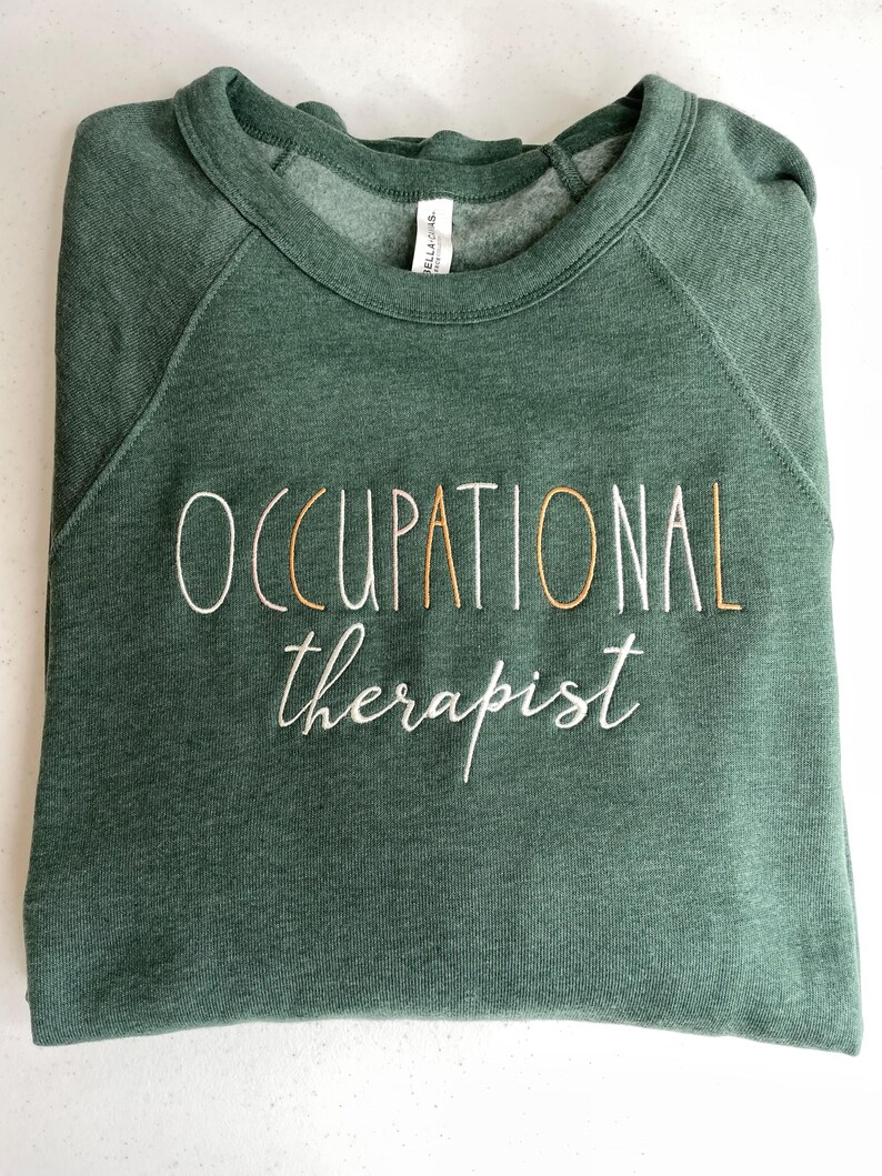 Może przedstawiać: Zielony sweter z haftem "OCCUPATIONAL therapist" w kolorze białym i złotym.