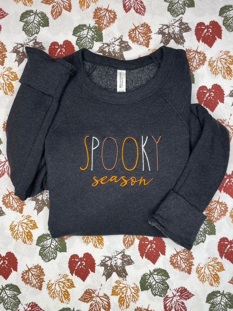 May include: Black sweatshirt with the text "SPOOKY season" in orange, white, and brown embroidery. The sweatshirt is on a white background with a pattern of brown, orange, and green leaves.