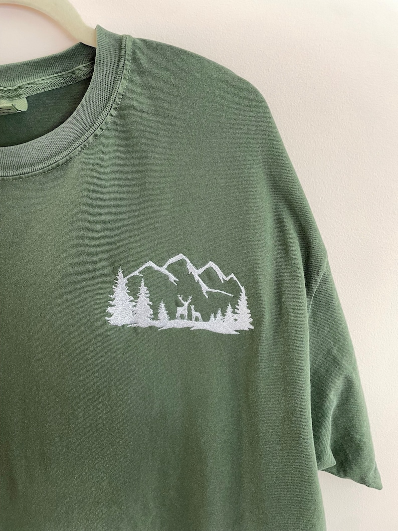 Pu&ograve; includere: Una maglietta verde con un disegno ricamato bianco di una catena montuosa con alberi e un cervo.