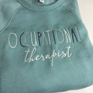 Może przedstawiać: Turkusowa bluza z haftem "Occupational therapist" wykonanym białą i czarną nicią.