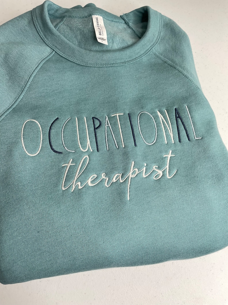 Może przedstawiać: Turkusowa bluza z haftem "Occupational therapist" wykonanym białą i czarną nicią.