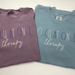 May include: Two t-shirts with the text "Occupational therapy" in different colors. The shirt on the left is a dark purple color and the shirt on the right is a light blue color.