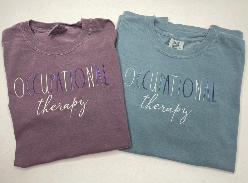 May include: Two t-shirts with the text "Occupational therapy" in different colors. The shirt on the left is a dark purple color and the shirt on the right is a light blue color.