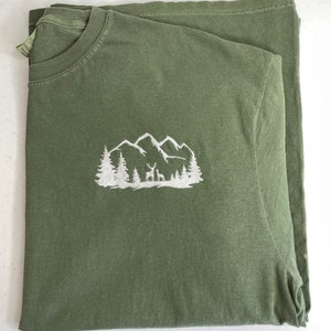 Pu&ograve; includere: Una maglietta verde con un disegno ricamato bianco di montagne, alberi e cervi.
