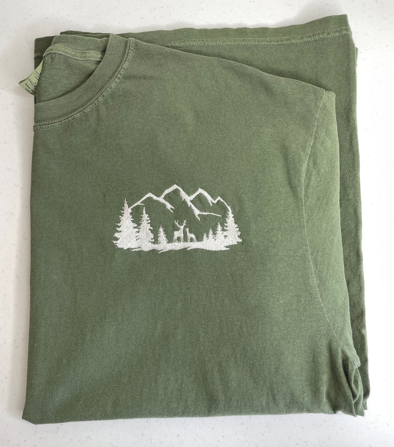 Pu&ograve; includere: Una maglietta verde con un disegno ricamato bianco di montagne, alberi e cervi.