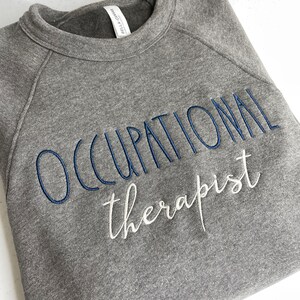 Może przedstawiać: Szara bluza z haftem "Occupational therapist" w kolorze niebieskim i białym.