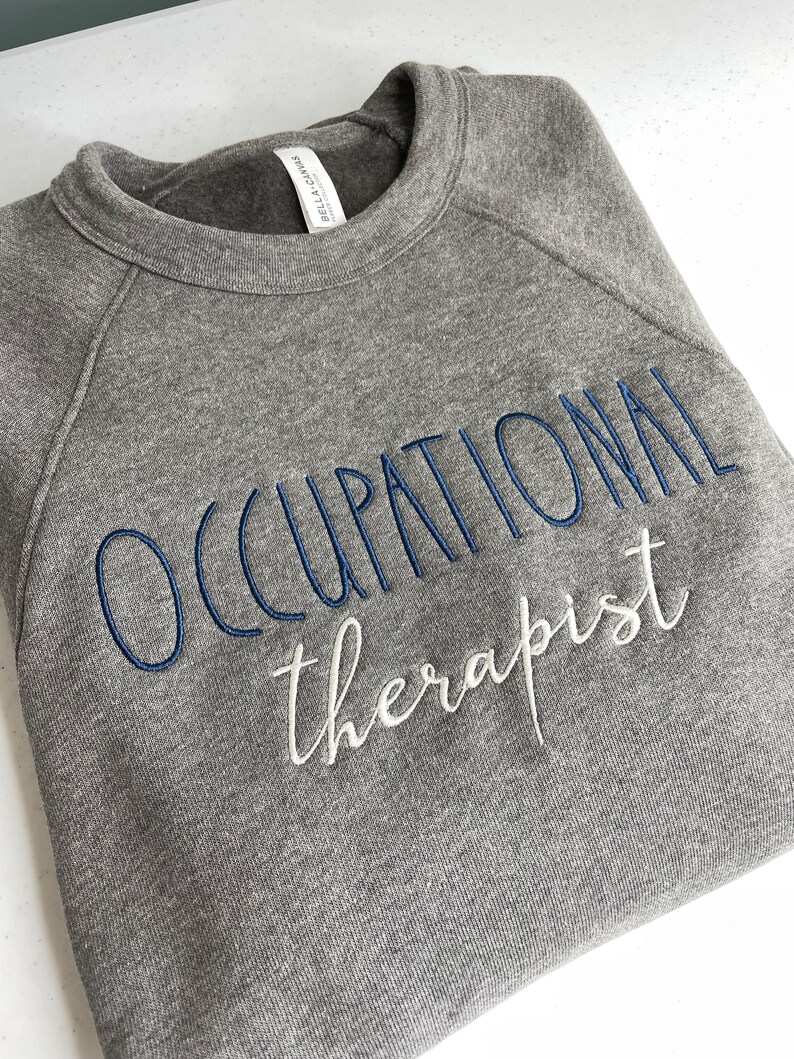 Może przedstawiać: Szara bluza z haftem "Occupational therapist" w kolorze niebieskim i białym.